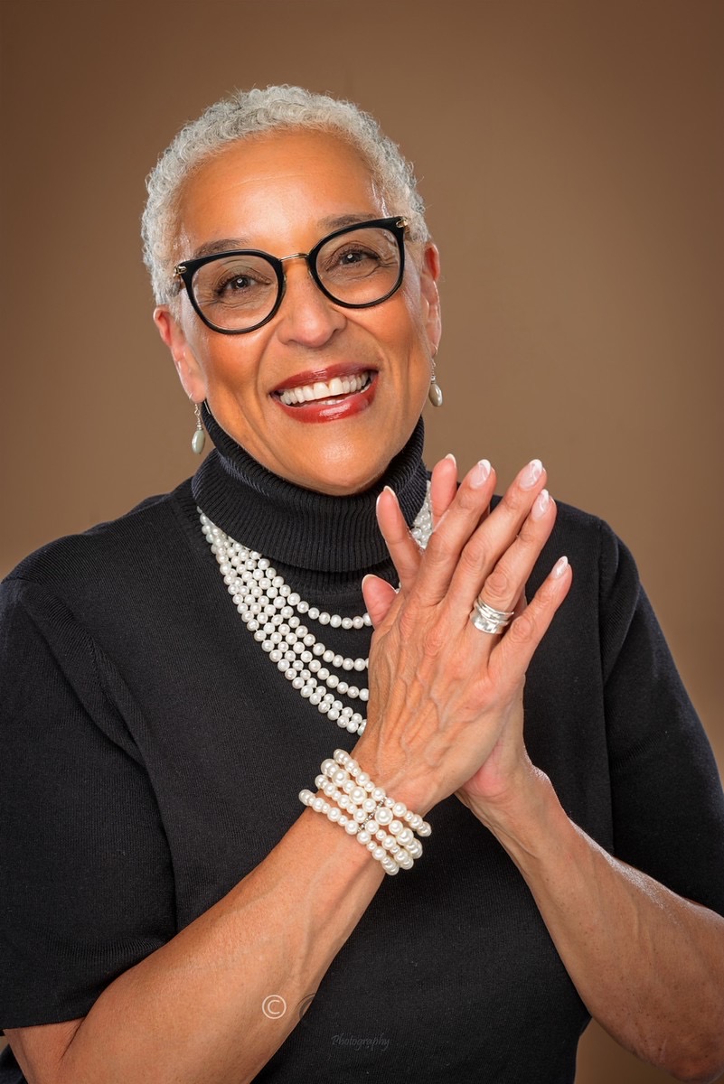 Rev. Dr. Zina Jacque smiling, hands pressed together at her heart, in a black turtleneck and pearls